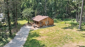 A wooden cabin with a porch, surrounded by trees and a grassy area.