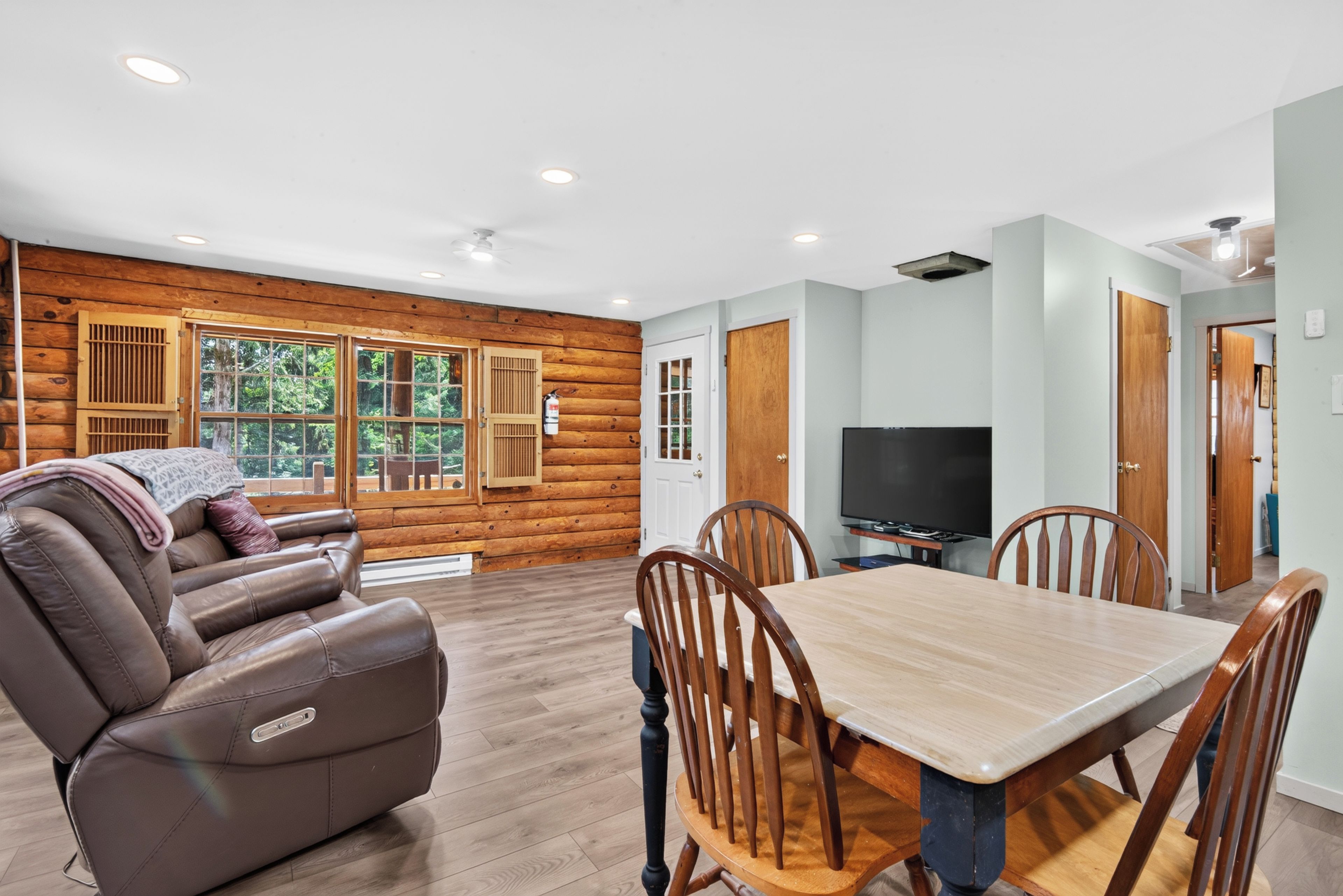 A log cabin interior with a leather sofa, dining table, chairs, a flat-screen TV, and a fireplace.