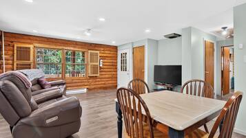 A log cabin interior with a leather sofa, dining table, chairs, a flat-screen TV, and a fireplace.