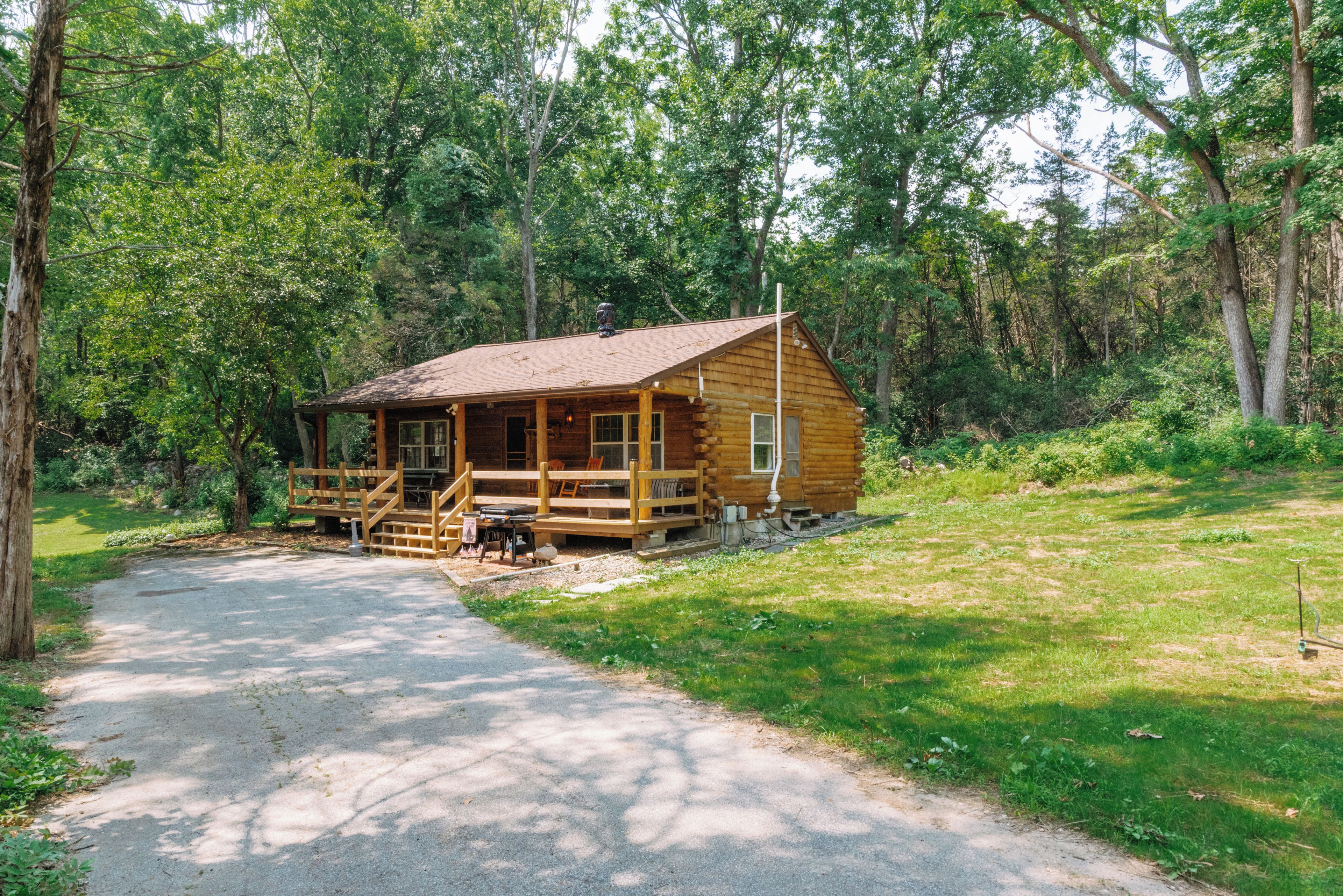 A wooden cabin with a porch, a gravel driveway, and a forested area.