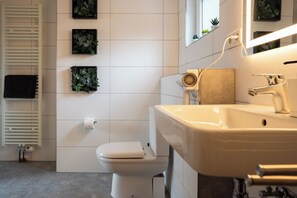 A modern bathroom with a white toilet, a beige sink, and a mirror above the sink. There are three framed plants on the wall, and a towel rack is visible. - Coal Street by Loftigo - House with a Large Garden (Übach-Palenberg)