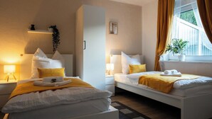 Hotellihuone, jossa on kaksi sänkyä, yöpöytä lampun kanssa, ikkuna verhoilla ja pieni hylly, jossa on kasvi.