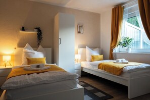 A hotel room with two beds, a bedside table with a lamp, a window with curtains, and a small shelf with a plant. - Coal Street by Loftigo - House with a Large Garden (Übach-Palenberg)