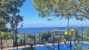 Un terrain de sport entouré d’un filet, bordé d’arbres et offrant une vue sur la mer.