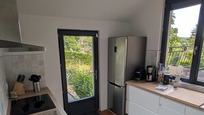 A modern kitchen with a built-in stove, a refrigerator, and a window with a view of greenery. - House for 8 people • Panoramic View of Lac du Causse • Rated ⭐⭐⭐⭐ • Air-conditioned (Lissac-sur-Couze)