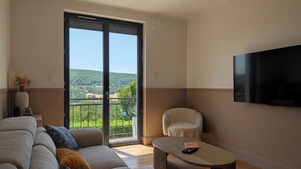 A living room with a sofa, a coffee table, a chair, a TV, and a sliding glass door leading to a balcony with a view of greenery. - House for 8 people • Panoramic View of Lac du Causse • Rated ⭐⭐⭐⭐ • Air-conditioned (Lissac-sur-Couze)