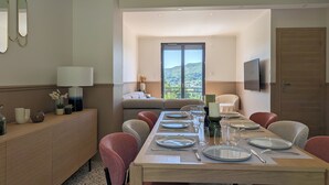 A dining area with a long table set for four, a sofa, a TV mounted on the wall, and a view of a mountainous landscape through a sliding door. - House for 8 people • Panoramic View of Lac du Causse • Rated ⭐⭐⭐⭐ • Air-conditioned (Lissac-sur-Couze)