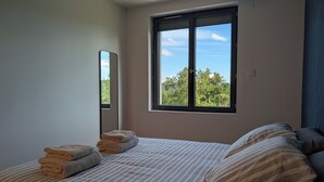 A bedroom with a large window, a bed with striped bedding, and a stack of folded towels. - House for 8 people • Panoramic View of Lac du Causse • Rated ⭐⭐⭐⭐ • Air-conditioned (Lissac-sur-Couze)
