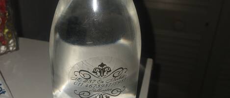 Una botella de vidrio transparente con tapón blanco, grabada con un logotipo y texto, sobre una superficie blanca.