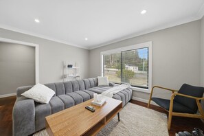 Ein modernes Wohnzimmer mit einem grauen Ecksofa, einem Couchtisch aus Holz, einem Holzsesseln und einem Fenster mit Jalousien.