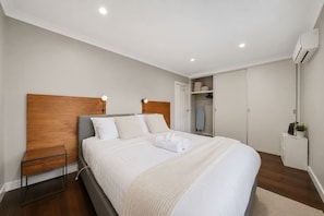Une chambre à coucher moderne avec une tête de lit en bois, un lit recouvert de linge de maison blanc, une table de chevet et une armoire.
