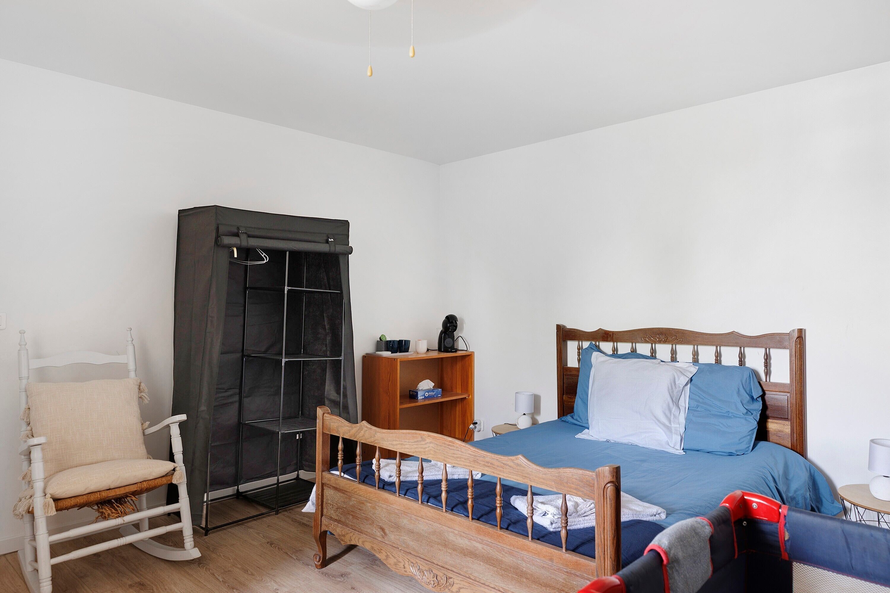 Een slaapkamer met een houten bed, een witte schommelstoel, een garderobe en een klein houten nachtkastje.