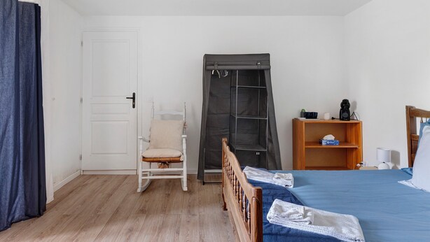 Een slaapkamer met een bed, een stoel, een kledingkast en een plank met spullen.