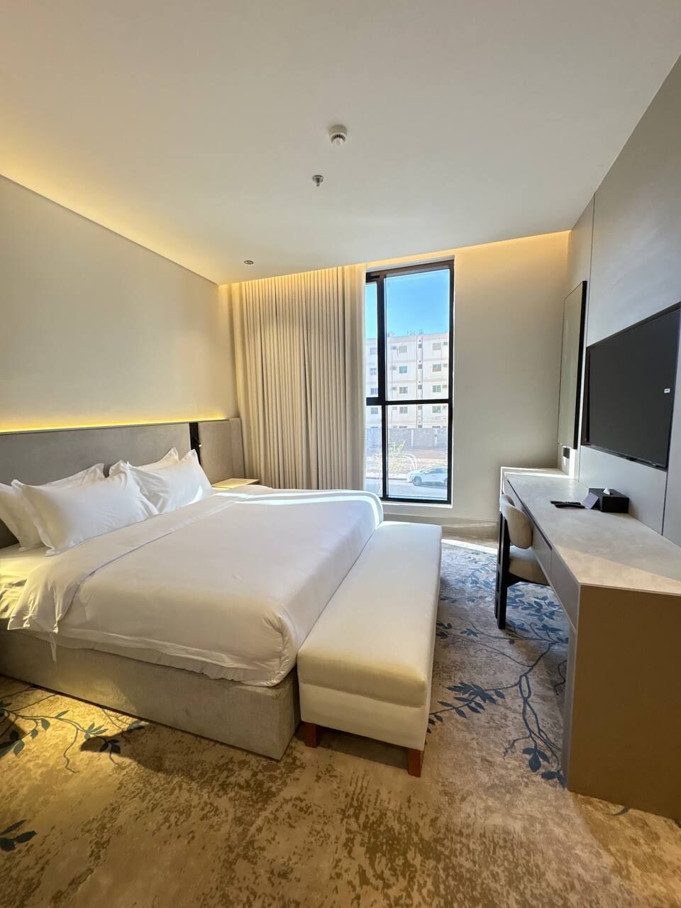 A hotel room with a bed, a desk, a chair, a large window, and a television.