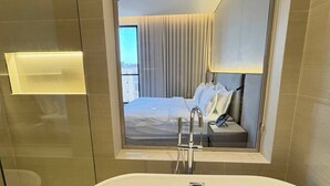 Um banheiro moderno com uma banheira grande, uma janela com vista para um quarto, e um espelho sobre a banheira.