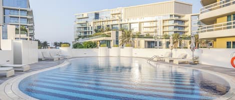 Una piscina con sillas de descanso, una zona cubierta y edificios modernos al fondo.