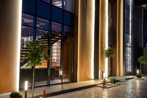 Fachada moderna de un edificio con amplios ventanales, una escalera y un acceso pavimentado con pequeños árboles.