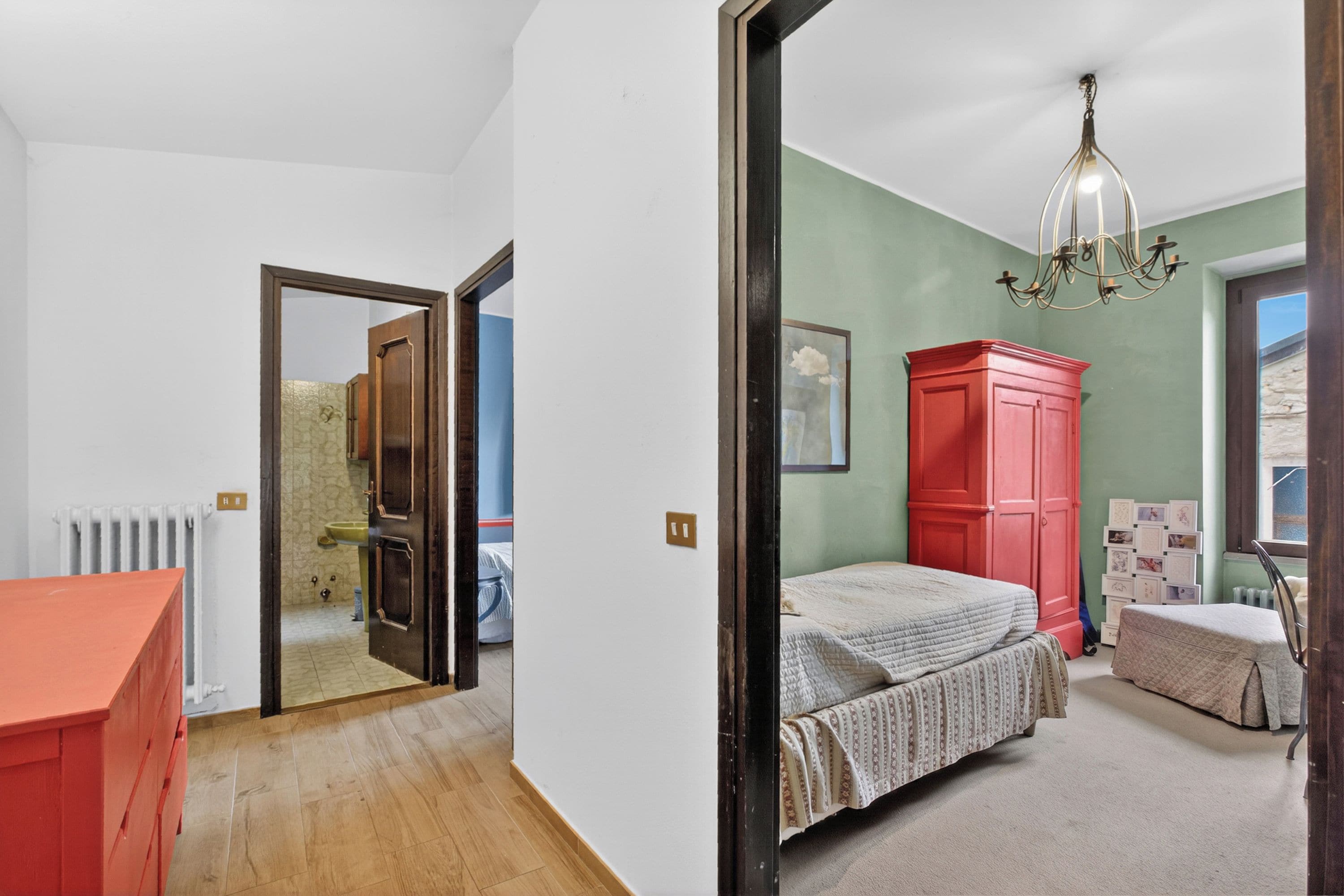 A bedroom with a bed, a red wardrobe, a chandelier, and a view of the outside.
