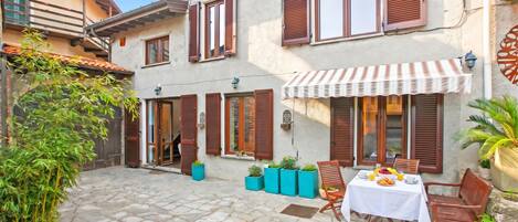 A courtyard with stone flooring, a table with chairs, potted plants, and a building with shutters.
