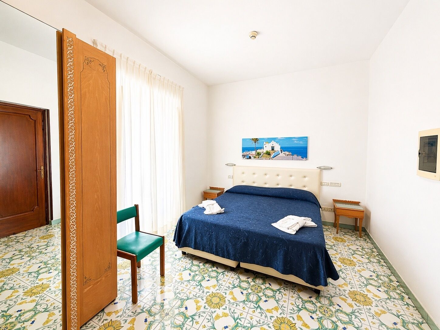 Une chambre à coucher avec un lit, une chaise, une fenêtre avec des rideaux, un tableau au mur et une porte.
