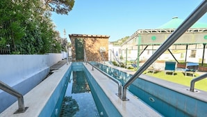 Una piscina con un canal revestido de piedra, un pequeño edificio con puerta verde y un área cubierta con sillas.