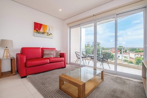 A modern living room with a red sofa, a glass coffee table, a painting, and a large sliding glass door leading to an outdoor balcony with a view of buildings and greenery. - Corporate Stays Edificio Pilar - N4A1 (Asunción)