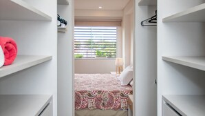 A narrow walk-through closet with open shelving, a bed with patterned bedding, and a window with blinds. - Corporate Stays Edificio Pilar - N4A1 (Asunción)