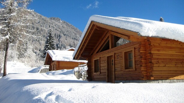 Een houten hut met een met sneeuw bedekte dak, omgeven door besneeuwde bomen en een berg op de achtergrond.