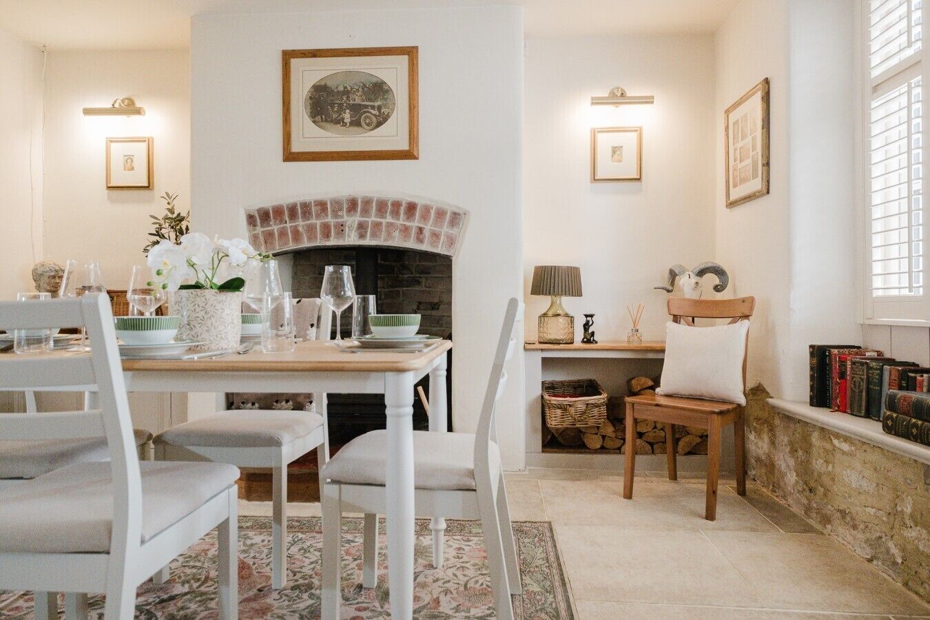 A dining area with a brick fireplace, white chairs, a table set for four, and framed pictures on the walls.