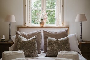 A bedroom with a bed, two nightstands, two lamps, a window with a view of greenery, and a vase with flowers. - Beautiful Cottage in Centre of Cirencester (Cirencester)