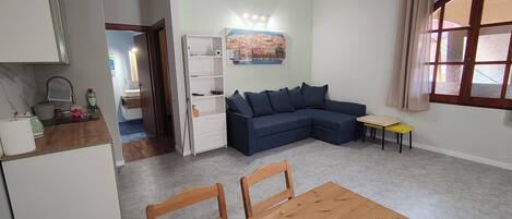Una sala de estar con sofá, una mesa de comedor con sillas, una zona de cocina y un ventanal con cortinas.