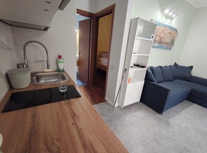 A compact living space with a kitchenette, a sofa, and a door leading to another room. - Cozy Apartment with Pool & Private Garden near Cagliari (La Maddalena)