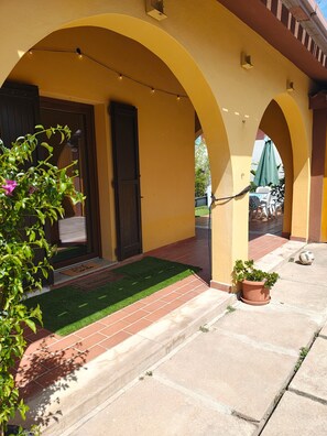 A building with yellow arches, a door, a small green lawn, and a potted plant. - Cozy Apartment with Pool & Private Garden near Cagliari (La Maddalena)