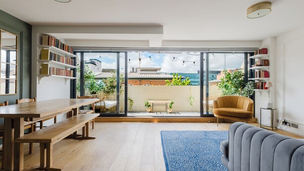 A modern living room with a large sliding glass door leading to a balcony, a wooden dining table, a sofa, and a bookshelf. - Refined 3BDR Retreat with Skyline Views (Greater London)