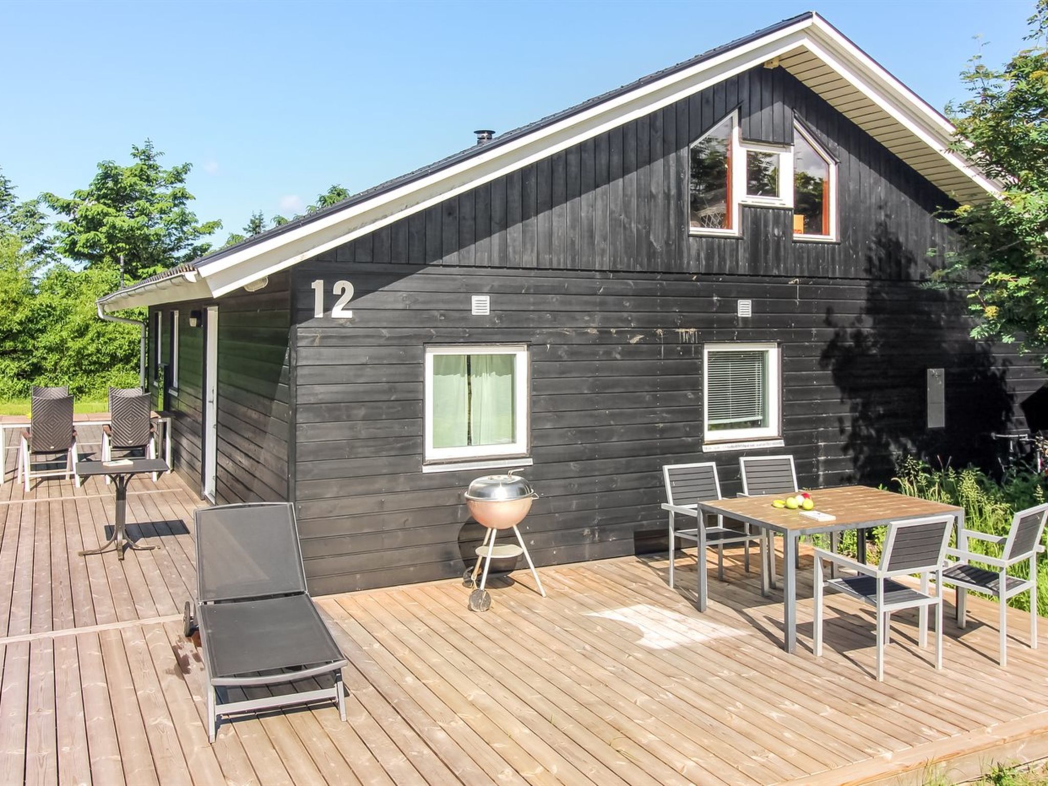 Una cabaña de madera negra con el número 12, una terraza amueblada y una parrilla.