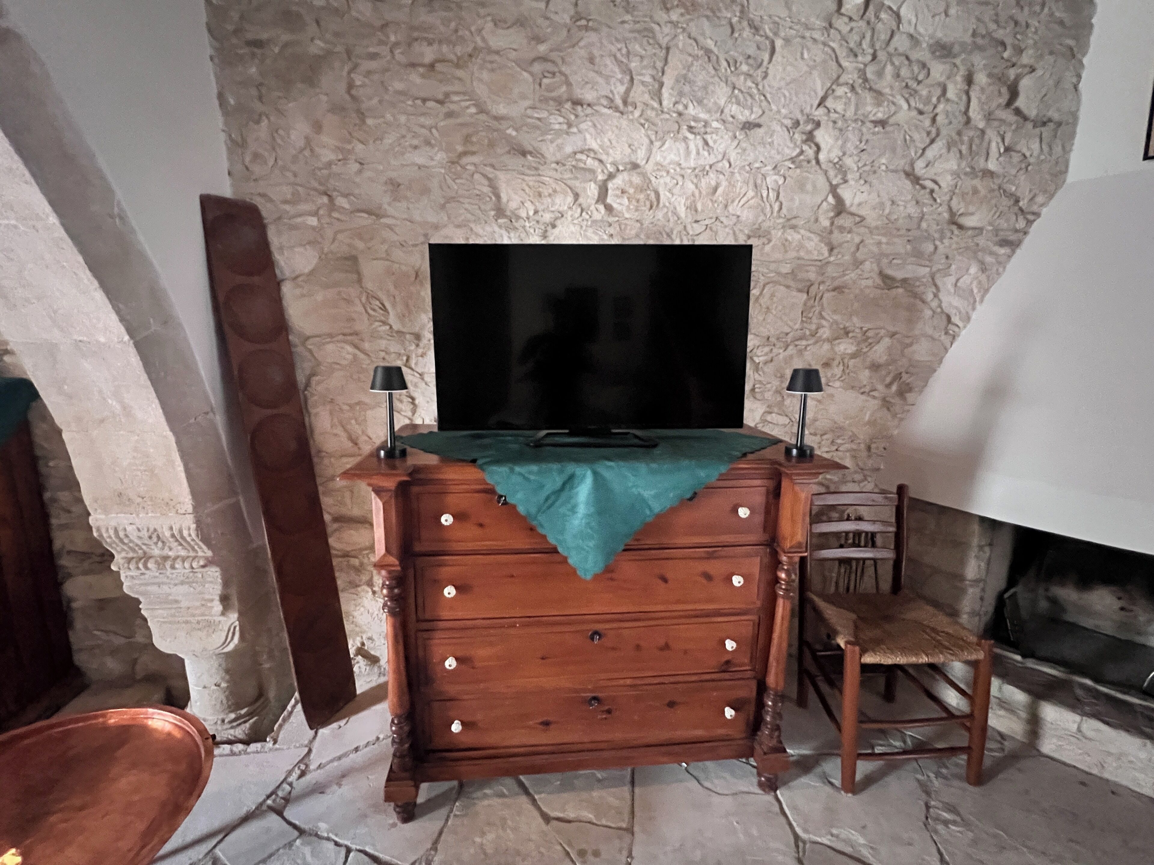 Un muro de piedra, un aparador de madera con cajones, un televisor de pantalla plana, dos lámparas, una silla y una chimenea.