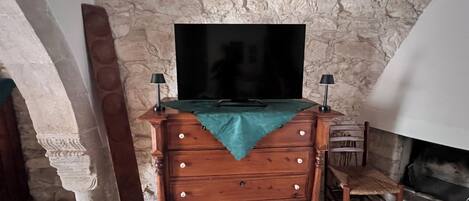 Un muro de piedra, un aparador de madera con cajones, un televisor de pantalla plana, dos lámparas, una silla y una chimenea.