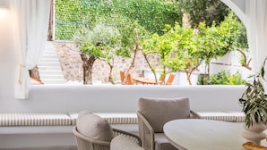 A room with a view of a garden through an arched window, a round table, and a sofa with striped cushions. - Nama Summer Retreat Mikra Mantinia Beachfront Gem (Kalamata)