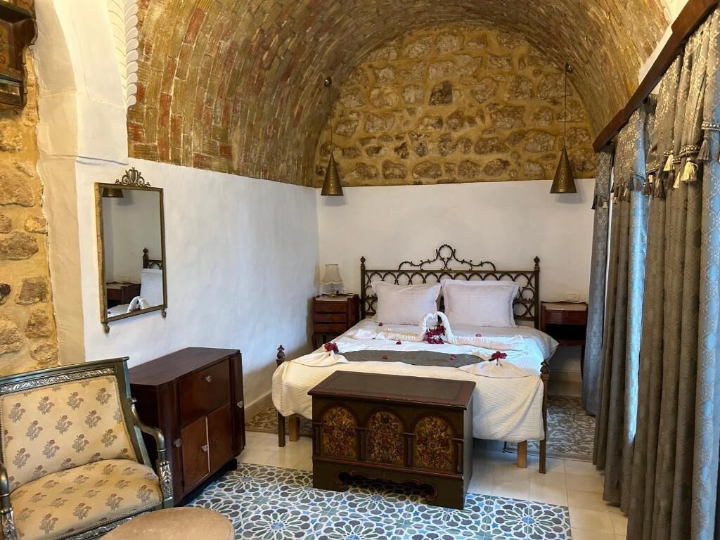 A room with a stone ceiling, a bed with a headboard, a nightstand, a dresser, a chair, and a patterned rug.