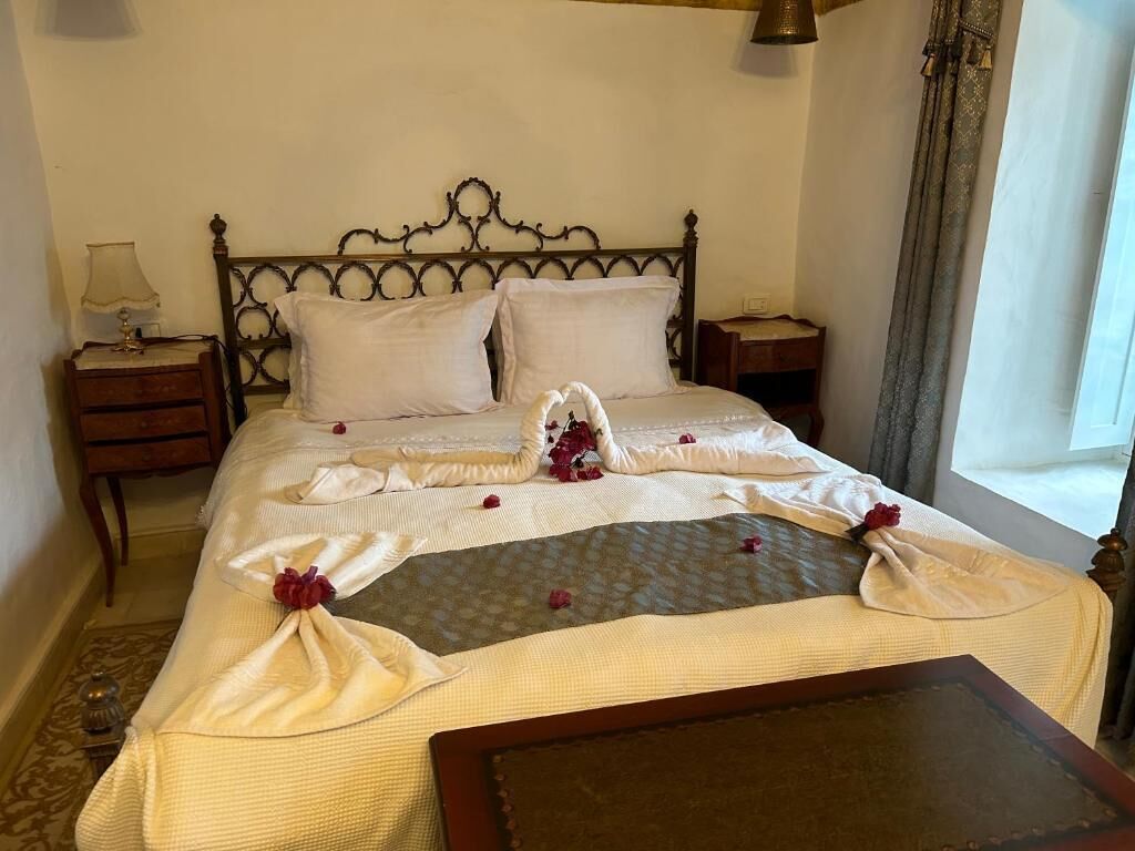 A neatly made bed with a decorative towel arrangement, a bedside table with a lamp, and a window with curtains.