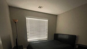 Ein Schlafzimmer mit Bett, Nachttisch, Lampe und einem Fenster mit Jalousien.