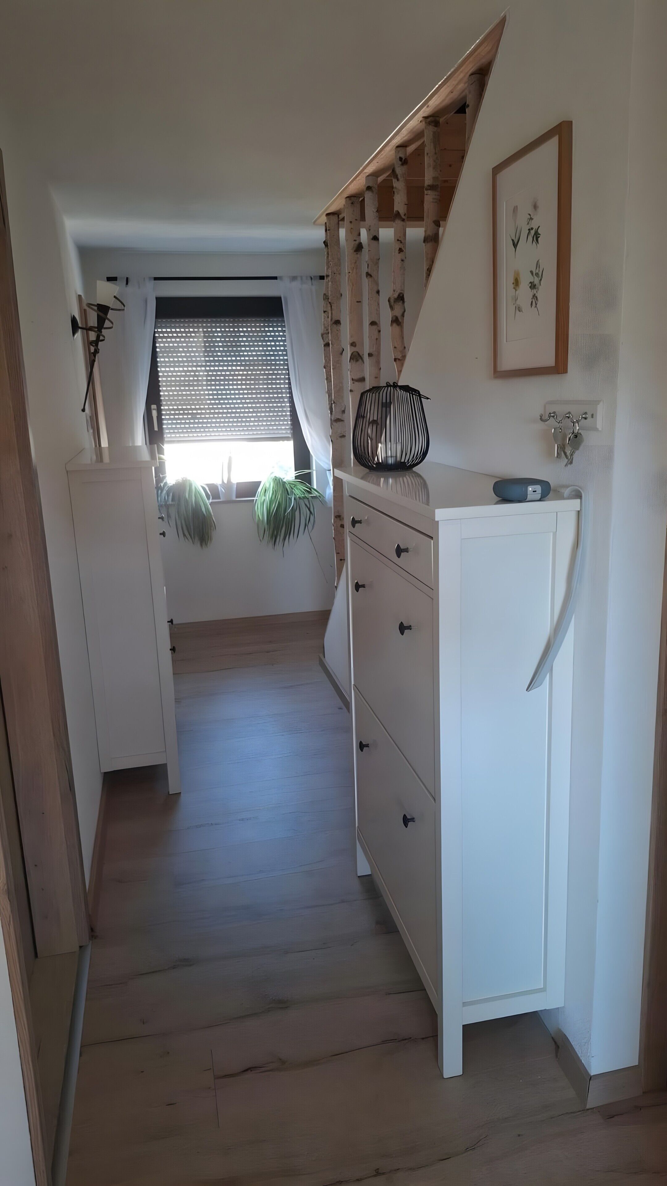 Ein schmaler Flur mit einer Holztreppe, einem weißen Schrank, einem kleinen Kühlschrank und einem Fenster mit Jalousien.