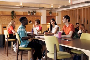 Eine Gruppe von Personen sitzt an einem langen Tisch in einem modernen Innenraum mit Holzvertäfelung und Pflanzen auf Regalen.