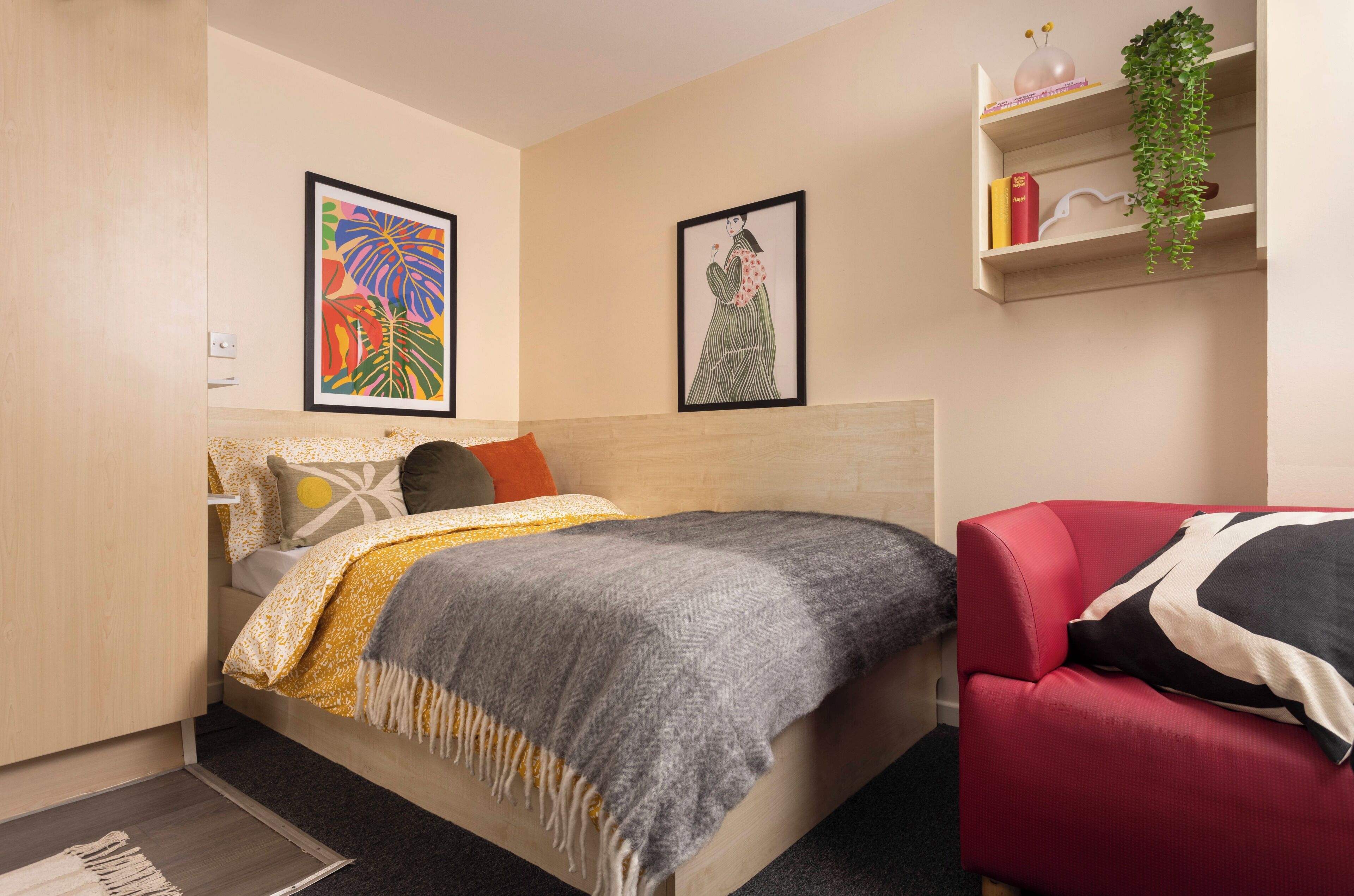 Ein Schlafzimmer mit einem Bett, einem roten Sessel, einem Regal mit Büchern und einer Pflanze sowie zwei gerahmten Kunstwerken an der Wand.