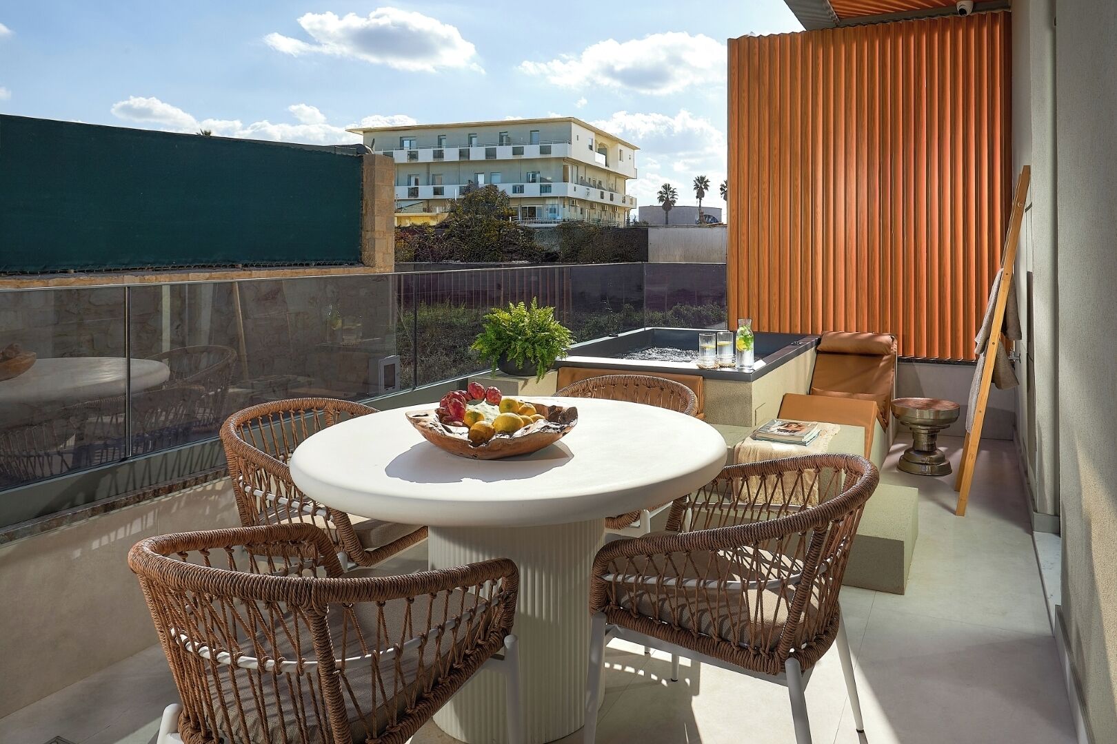 Un balcon avec des fauteuils en osier, une table ronde sur laquelle est posé un panier de fruits, un jacuzzi et une vue sur les bâtiments.