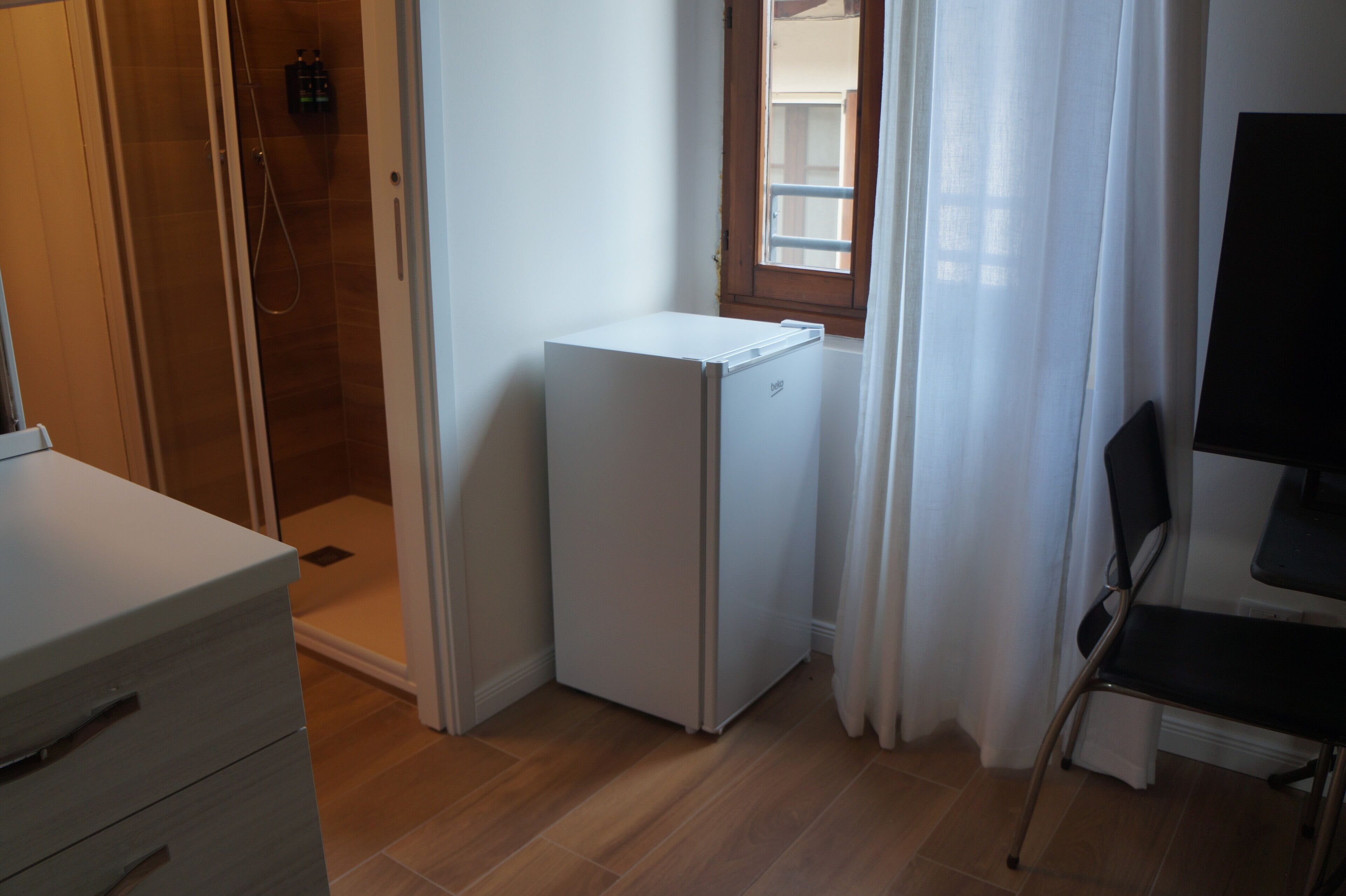 A compact room with a shower, a small white refrigerator, a chair, and a desk.