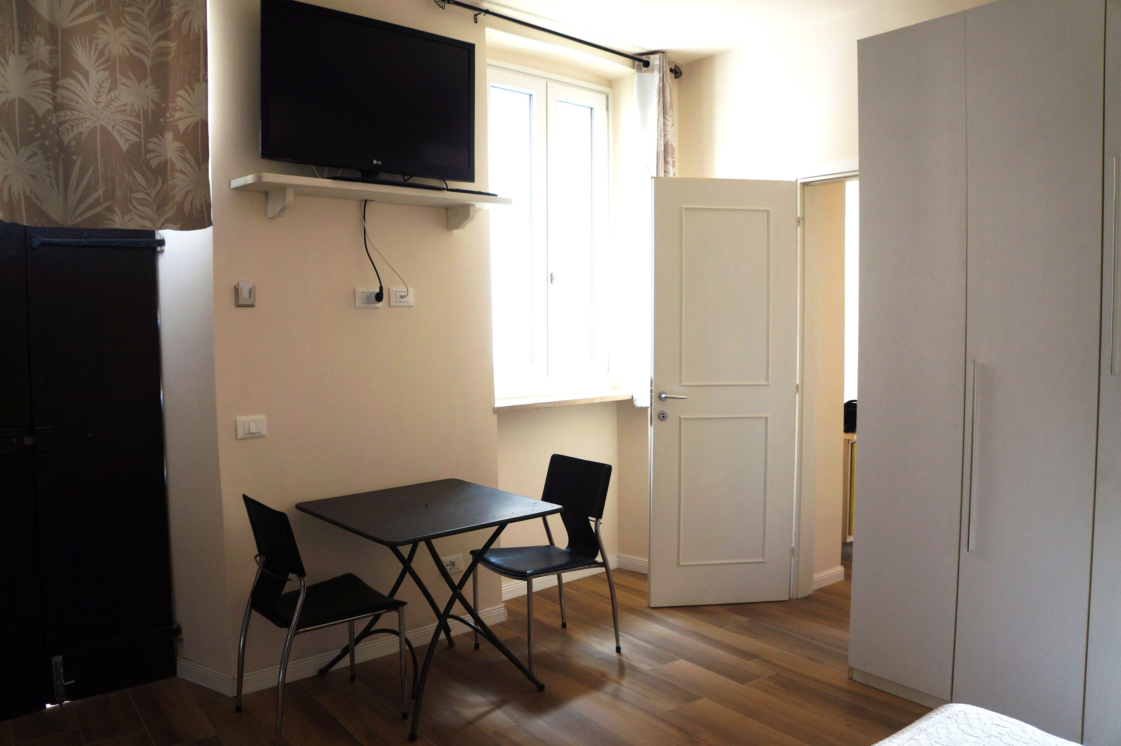 A room with a TV mounted on the wall, a dining table with chairs, a white door, and a wardrobe.