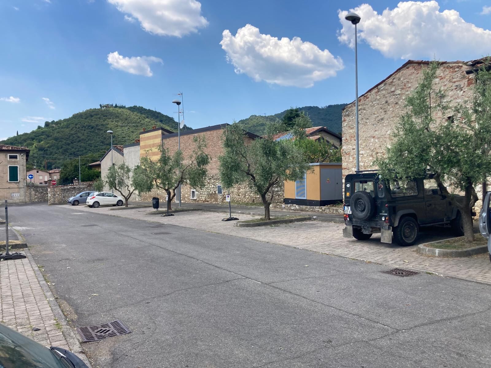 A quiet street with parked cars and a black SUV, stone buildings, and a hill in the background.