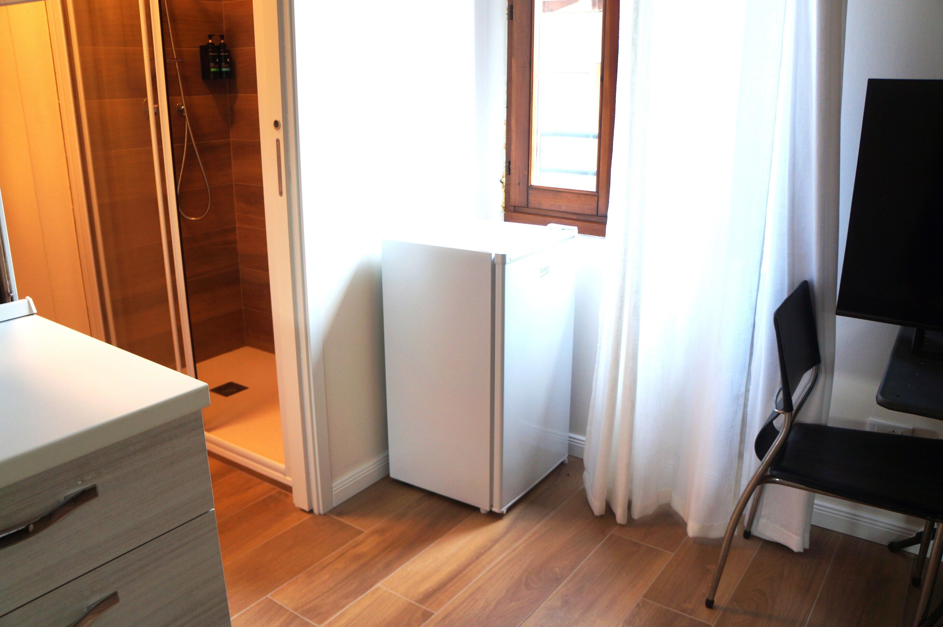 A small room with a shower, a refrigerator, a chair, and a window with curtains.