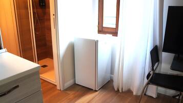 A small room with a shower, a refrigerator, a chair, and a window with curtains.